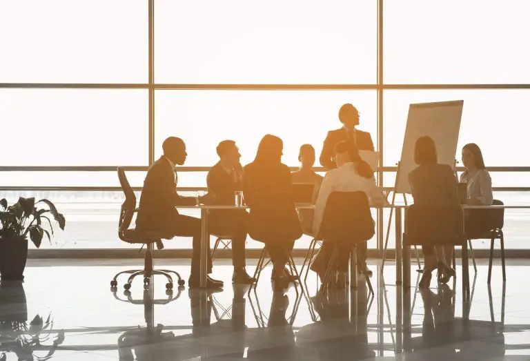 Business team in a modern office engaged in a strategic discussion, with a leader presenting at a whiteboard. Sunlight fills the room, highlighting collaboration.