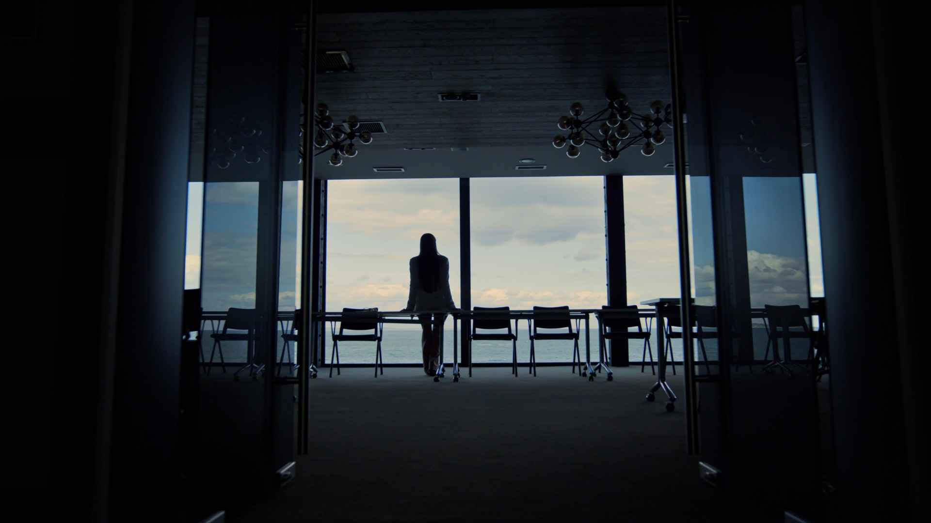 Femme dans une salle de conférence regardant l’horizon par une grande fenêtre sur la mer.